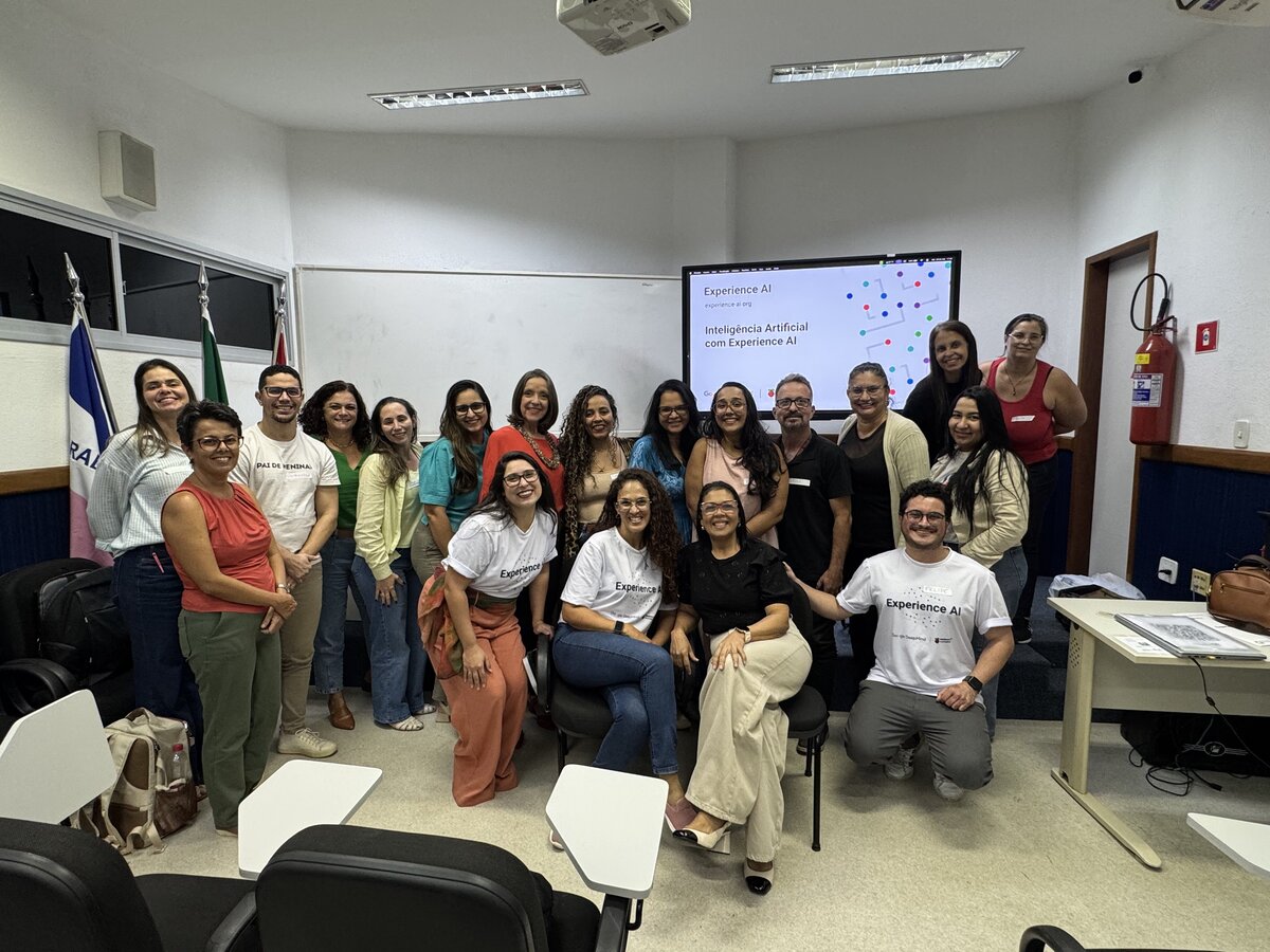 Grupo de pessoas reunidas em sala de aula. Ao fundo, uma tela exibe: 'Experience AI, experience-ai.org, Inteligência Artificial com Experience AI'. Alguns participantes vestem camisetas do projeto.