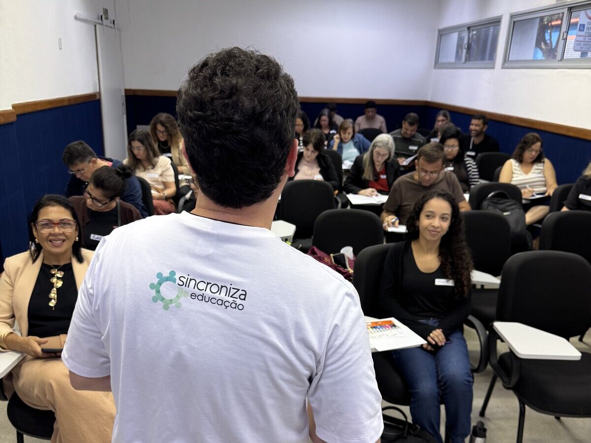 Homem de costas, vestindo camiseta branca com o logotipo 'Sincroniza Educação', fala para uma sala cheia de pessoas sentadas em carteiras universitárias prestando atenção.