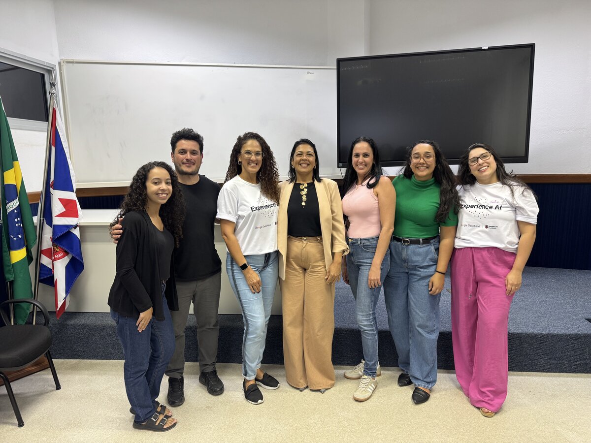 Sete pessoas posam sorrindo lado a lado em uma sala de aula à frente de um quadro branco e uma TV desligada. Duas mulheres vestem camisetas brancas do projeto 'Experience AI'.