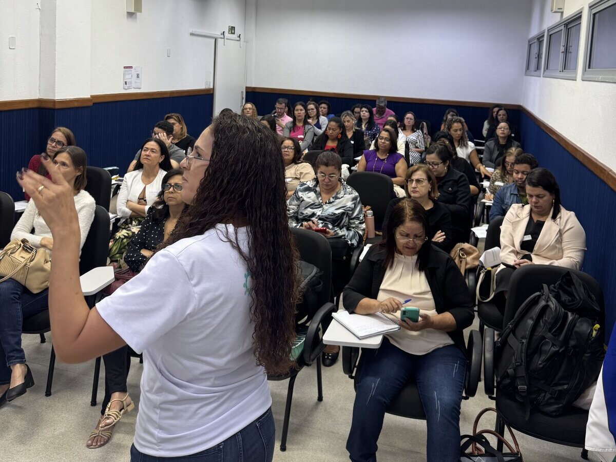 Mulher de perfil e cabelos cacheados compridos gesticula com a mão esquerda enquanto fala para uma plateia de pessoas sentadas em uma sala de aula de formato retangular.