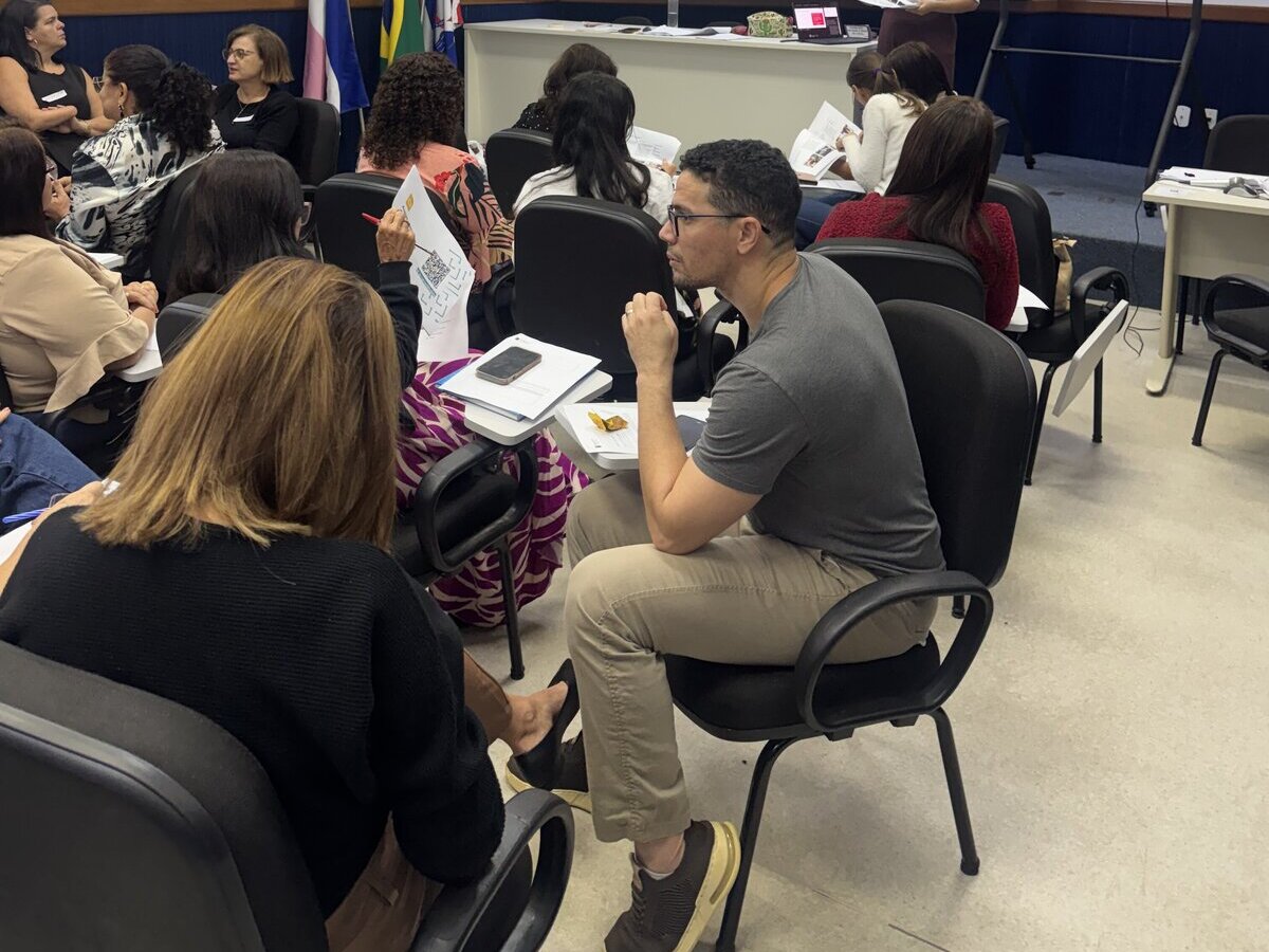 Homem de óculos e camiseta cinza, sentado de perfil em uma carteira acadêmica, conversa com uma colega à sua frente. Ao redor, outras pessoas participam da atividade na sala.