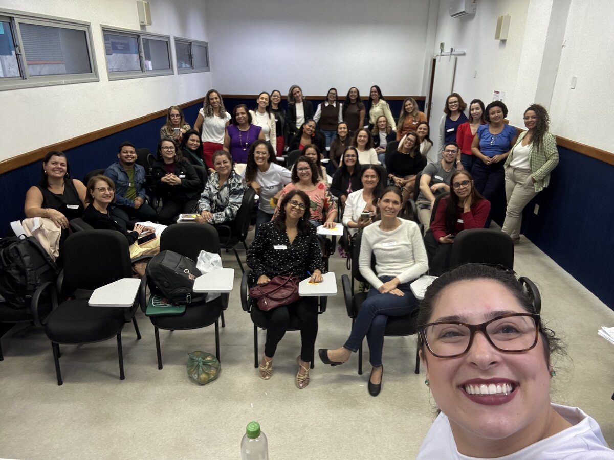 Mulher sorridente em primeiro plano tira uma foto estilo selfie mostrando um grande grupo de pessoas sentadas em carteiras acadêmicas organizadas em fileiras dentro de uma sala de aula.