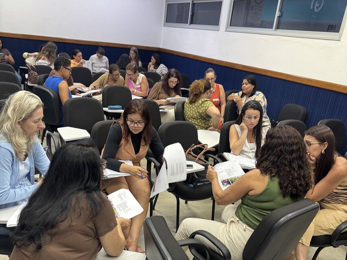 Participantes divididos em pequenos grupos de discussão dentro de uma sala de aula. Eles estão sentados em carteiras acadêmicas universitárias voltadas umas para as outras, lendo e analisando papéis impressos.