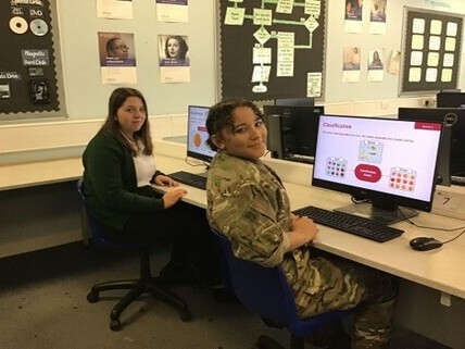 Duas jovens sentadas em um laboratório de informática sorriem para a câmera. À direita, um monitor exibe um slide didático intitulado 'Classification'.