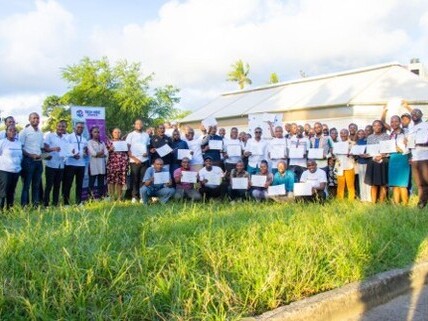 Grande grupo de pessoas reunido ao ar livre em uma área gramada, posando para foto coletiva. Quase todos os participantes estão segurando folhas de papel branco ou certificados à frente do corpo.