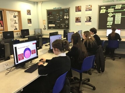 Estudantes sentados de costas e de perfil em uma bancada de laboratório de informática. Em primeiro plano, o monitor exibe um slide intitulado 'How do models learn?'.